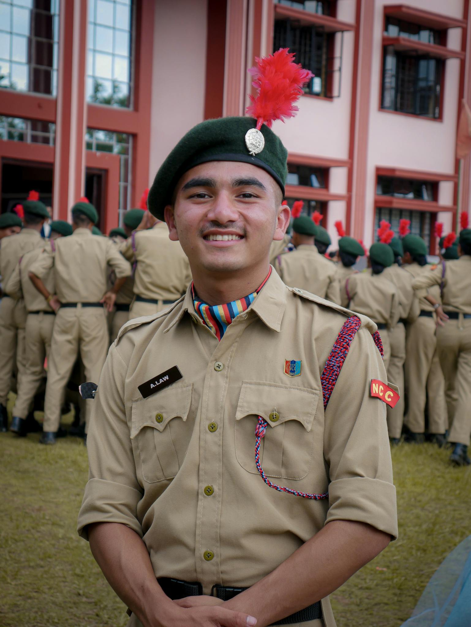 Smiling young male cadet in uniform during NCC training, standing outdoors with fellow cadets.